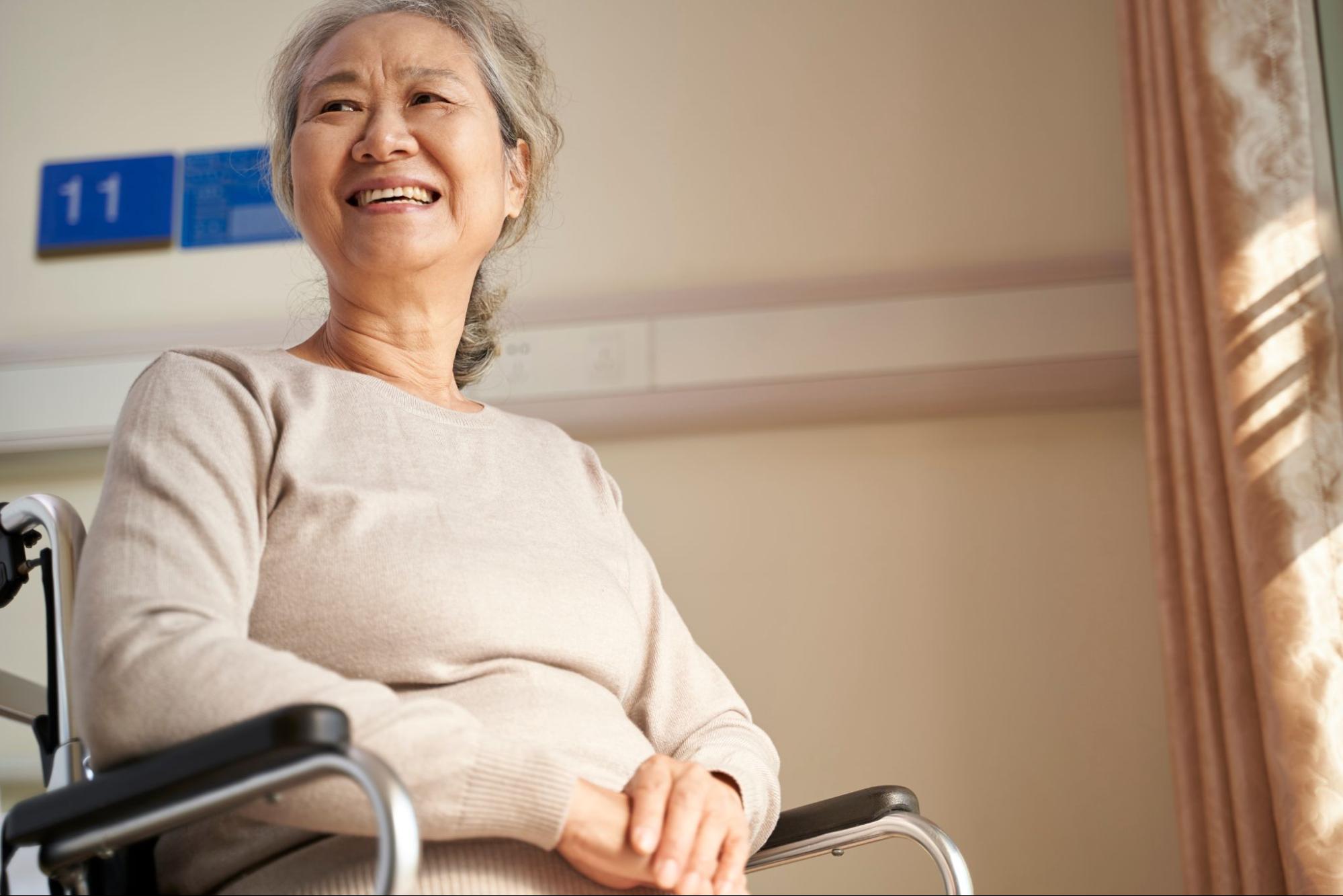 An elderly Asian woman with grey hair smiling warmly while sitting in a wheelchair, indoors by a window with sunlight streaming through, and a blue room number sign in the background.