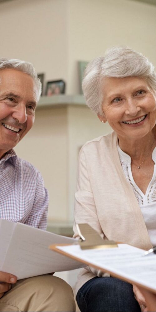 An elderly couple smiling warmly at someone off-camera while sitting on a couch, with the man holding papers and the woman beside him looking pleased. They appear to be in a discussion with a person holding a clipboard, suggesting a friendly consultation or meeting at home.