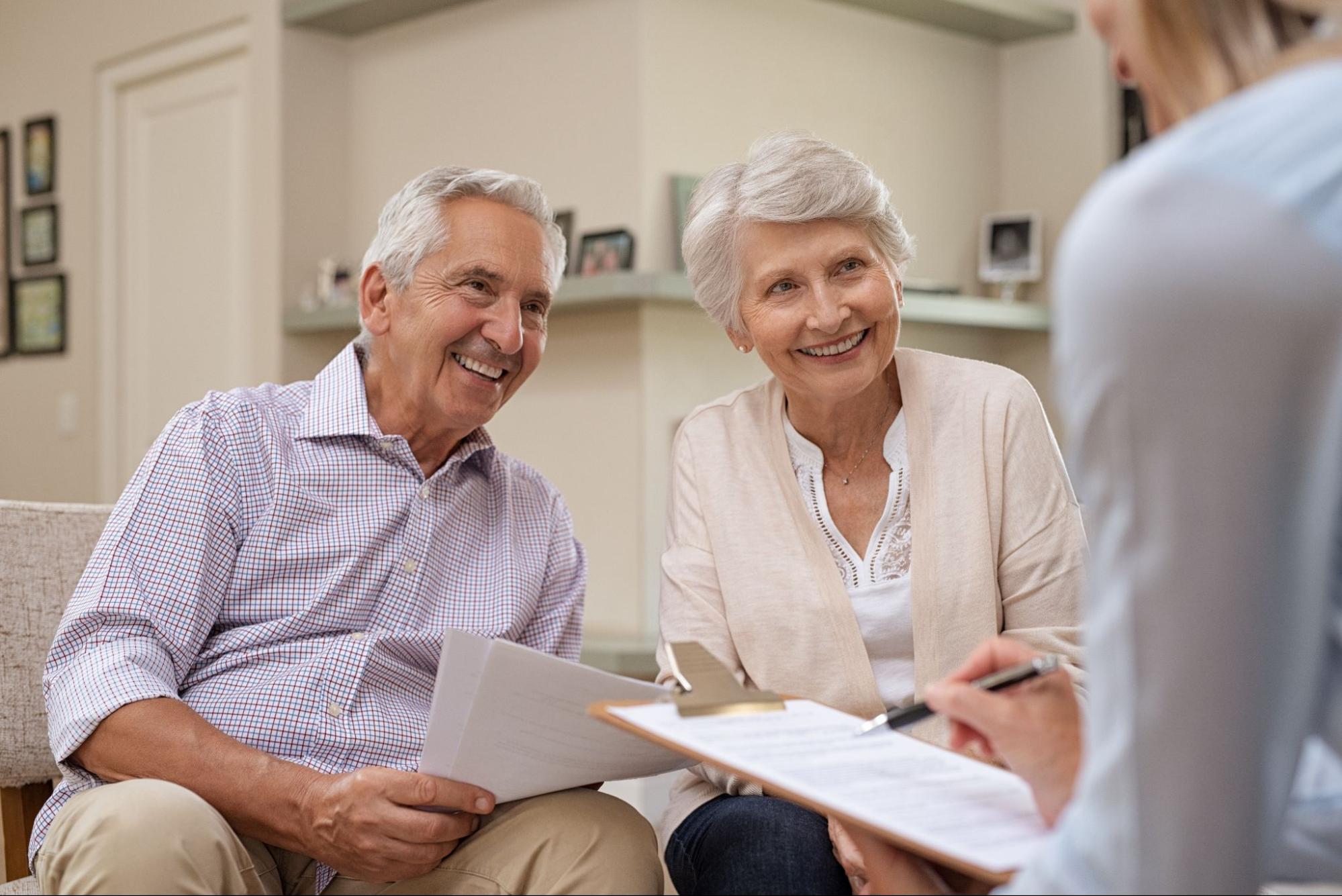 An elderly couple smiling warmly at someone off-camera while sitting on a couch, with the man holding papers and the woman beside him looking pleased. They appear to be in a discussion with a person holding a clipboard, suggesting a friendly consultation or meeting at home.